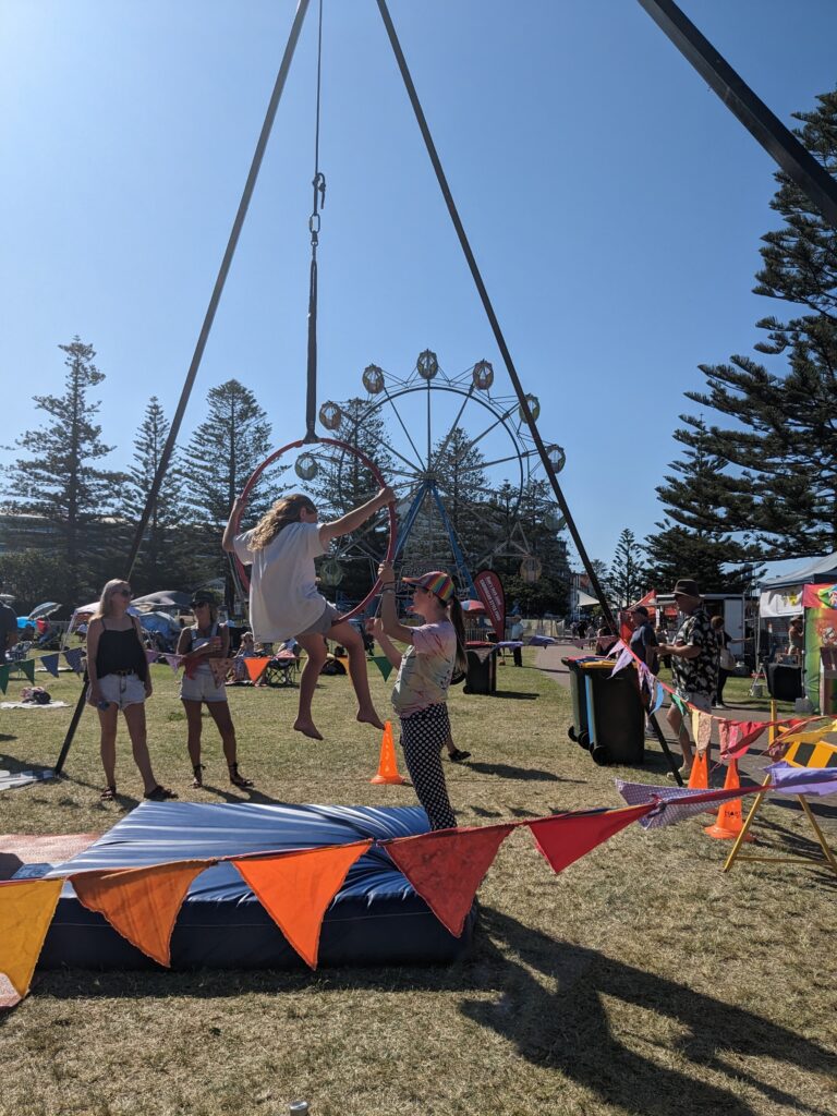 A child sitting on a Lyra aerial hoop, with an instructor providing support at Girrakool Blues festival in the Roundabout Circus play space.