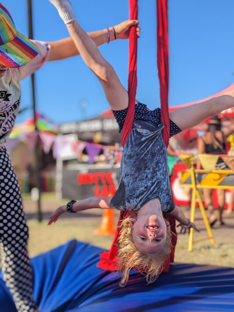 A child in a circus playspace hangs upside down on a vibrant red silk, supported by a trained instructor.